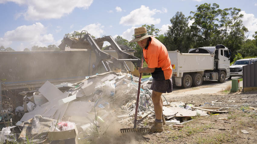 Worker raking up drywall scraps and construction waste on a building site, with a loader and dump truck behind