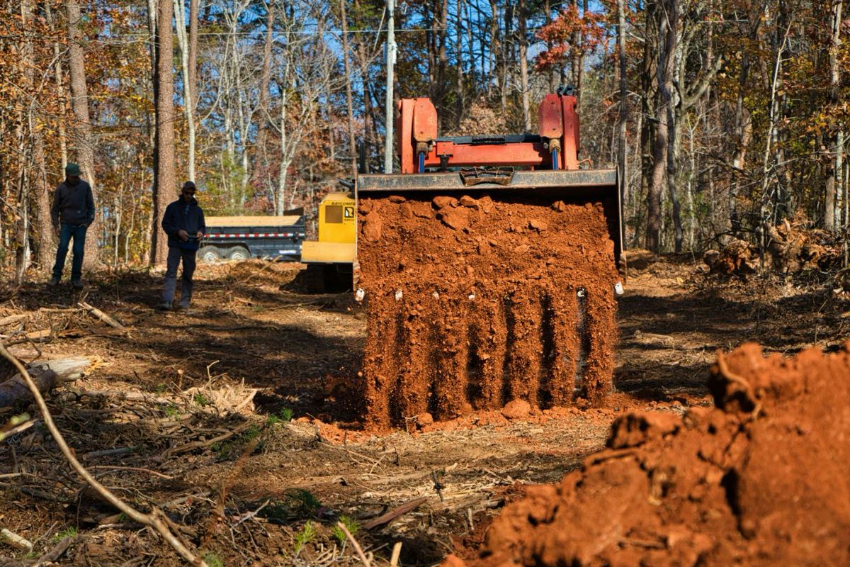 Track loader dumping a bucket of red dirt on a wooded lot being cleared, with crew and equipment behind